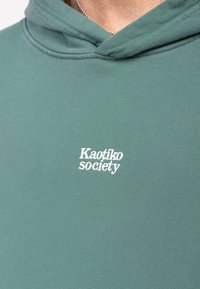Grön hoodie med vitt broderat "Kaotiko society"-logotyp på bröstet; tillverkad av mjukt, strukturerat tyg; har en huva och en avslappnad passform.