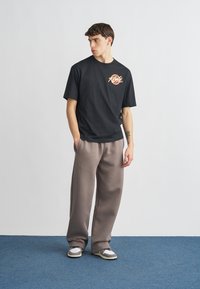 Melna T-krekla ar grafisku zīmējumu uz krūtīm, kopā ar vaļīgām pelēkām biksēm un sporta apaviem. Minimālistisks dizains ar atslābušu pieguļošu formu.