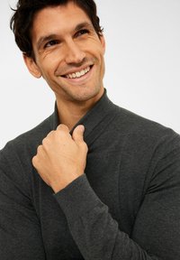 Dunkelgrauer Pullover mit hohem, geripptem Kragen. Weiche Textur, figurbetontes Design. Das Model lächelt und hält den Kragen mit einer Hand.