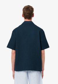 Μπλε ναυτικού κοντομάνικο πουκάμισο κατασκευασμένο από υφαντό ύφασμα, με γιακά και χαλαρή εφαρμογή, ορατές ραφές στο στρίφωμα.