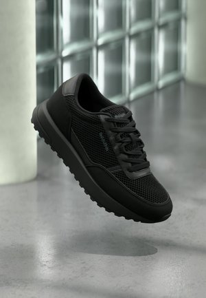 Schwarzer Sportsneaker mit Mesh- und Lederdetails, dicke Sohle und Schnürsenkel, schwebt über einem polierten Betonboden in der Nähe einer weißen Säule.