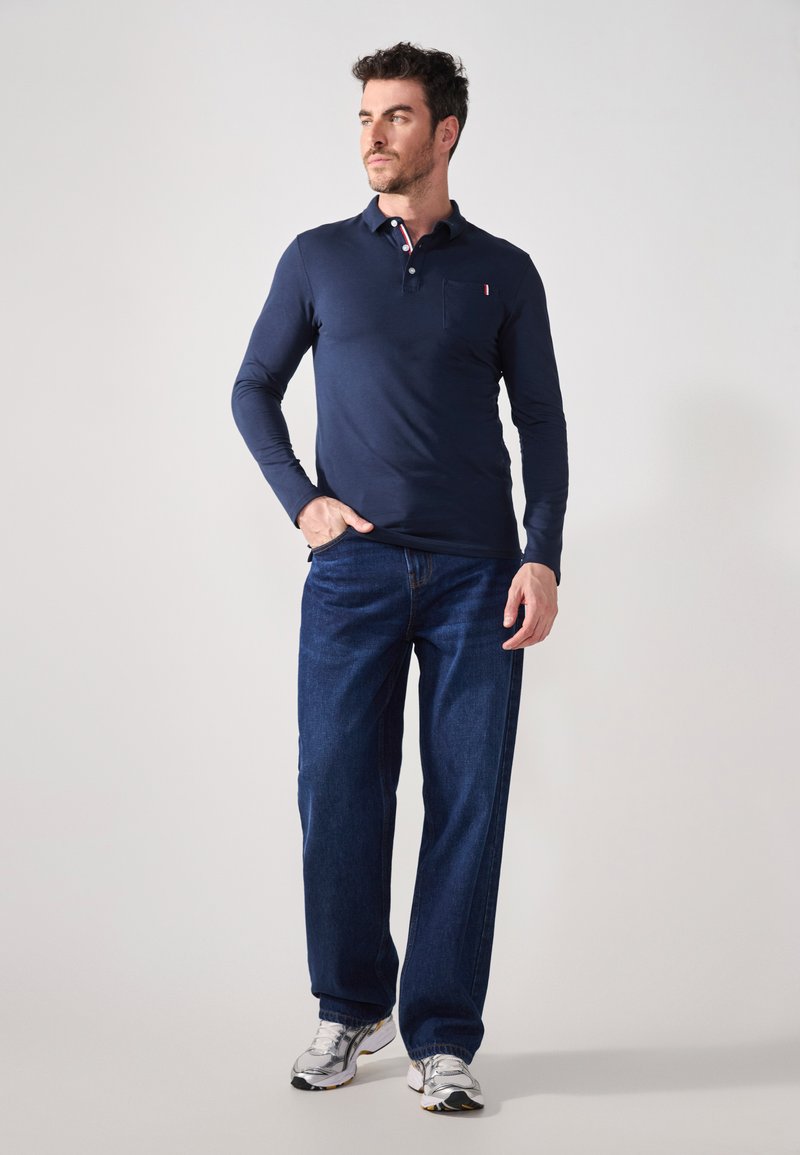 Marineblå langermet polo skjorte med krage og brystlomme, kombinert med vide blå jeans og grå joggesko.