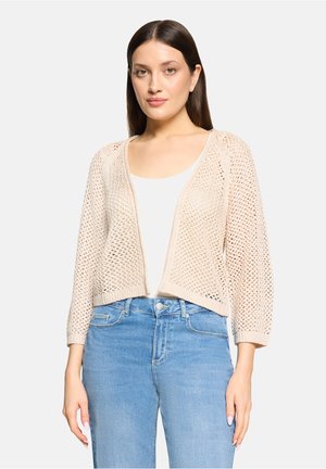 Femme portant un cardigan beige en maille ajourée sur un haut blanc et un jean taille haute bleu clair, debout devant un fond uni.