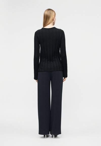 Femme aux longs cheveux blonds portant un pull côtelé noir et un pantalon large bleu marine, debout dos à un fond uni.