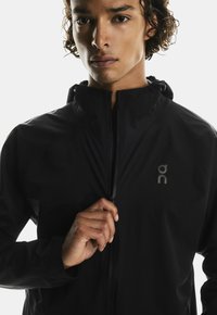 Giacca leggera nera con colletto alto e chiusura a zip. Presenta un piccolo logo sul petto e una texture liscia.