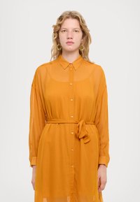 Rochie dintr-un material portocaliu transparent, cu guler, mâneci lungi și o curea legată la talie, având închidere cu nasturi și o siluetă fluidă.