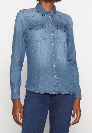 Kvinde iført en tætsiddende lyseblå denimskjorte med brystlommer og lange ærmer, kombineret med mørkeblå jeans.