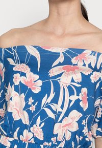 Haut bleu à fleurs avec épaules dénudées, présentant des motifs de fleurs roses et blanches, une taille froncée et une texture de tissu douce.