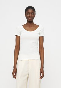 Woman with dark skin and hair pulled back wearing a short-sleeve white scoop-neck top and cream wide-leg trousers, standing against a plain background.