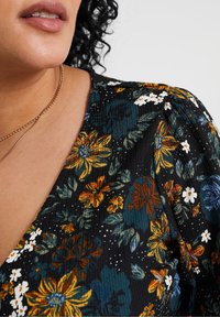 Schwarze Bluse mit floralen Motiven, die farbenfrohe Blumen in Gelb, Blau und Weiß zeigt. Sie besteht aus strukturiertem Stoff und hat puffige Ärmel. Eine Halskette mit goldenen Akzenten ist sichtbar.