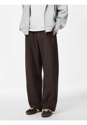 Osoba ubrana w luźne, ciemnobrązowe spodnie, ciemnobrązowe trampki z białymi sznurowadłami, białą koszulę i jasnoszary rozpinany kaptur. Ręce w kieszeniach.