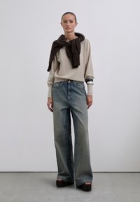 Mujer con un suéter beige y un suéter marrón oscuro sobre los hombros, jeans de pierna ancha y deslavados, sandalias de plataforma y pulseras de plata.