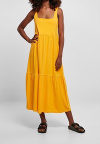 Żółta bezrękawnikowa sukienka midi z kwadratowym dekoltem, marszczonym gorsetem i falbaniastym dołem. Wykonana z miękkiego, lekkiego materiału, noszona z czarnymi sandałami.