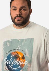 Una camiseta de color crema con un gráfico de palmeras y un atardecer, con "California" en texto en negrita. Manga corta, cuello redondo.
