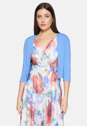 Frau trägt ein ärmelloses, geblümtes Kleid mit roten, blauen und grünen Mustern, kombiniert mit einer hellblauen, kurzen Strickjacke.