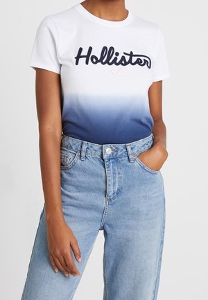 Osoba ubrana w biało-niebieską koszulkę ombre Hollister włożoną do jasnoniebieskich dżinsów z wysokim stanem, z jedną ręką w kieszeni.