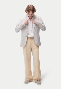 Veste de costume gris clair avec deux poches avant, fermeture à boutons, portée sur une chemise blanche à boutons. Pantalon beige à jambes larges et chaussures beiges.