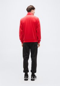 Κόκκινο αθλητικό μπουφάν με ψηλό γιακά, λείο υλικό και ελαστική κάτω ραφή, συνδυασμένο με μαύρα παντελόνια τζόγκινγκ και μαύρα αθλητικά παπούτσια.