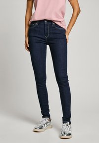 Sötét farmer skinny farmernadrág közepes magas derékkal, diszkrét varrásokkal és öt zsebes kialakítással, fémes sportcipővel és rózsaszín felsővel párosítva.