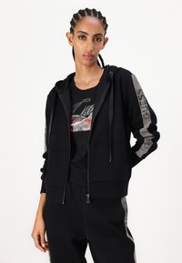Sudadera negra con cremallera y cordones, con acentos metálicos en las mangas, combinada con una camiseta gráfica negra. Tela suave, corte relajado.