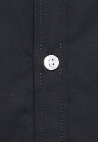 Camicia di tessuto nero con una superficie liscia, dotata di un bottone bianco sulla placca fissato da cuciture nere.