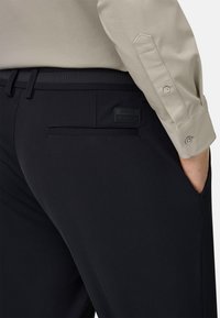 Schwarze, maßgeschneiderte Hose mit glatter Textur, ausgestattet mit einer Gesäßtasche und einem dezenten Logopatch. Kombiniert mit einem beigen Langarmhemd.