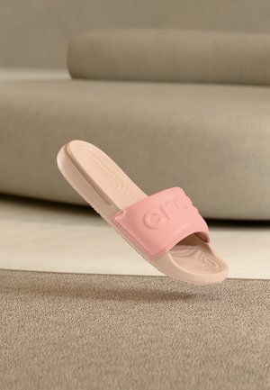 Sandale slide rose clair avec semelle texturée et bride avec logo embossé, flottant au-dessus d'un tapis beige dans une pièce doucement éclairée.