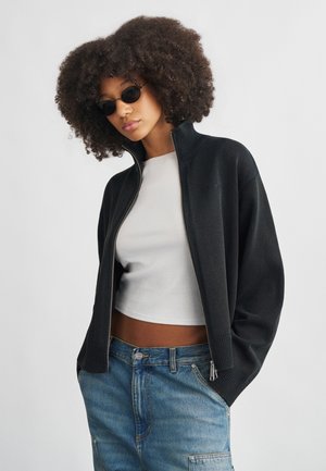 Cardigan noir à fermeture éclair avec texture côtelée, col montant, porté sur un crop top blanc, associé à un pantalon en denim bleu. Lunettes de soleil rondes noires portées.