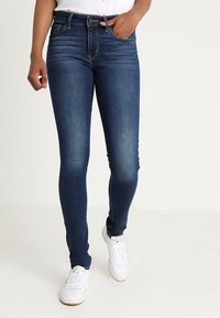Vaqueros ajustados de denim azul oscuro con un patrón deslavado, cintura alta y detalles sutiles en los bolsillos traseros, combinados con zapatillas blancas.