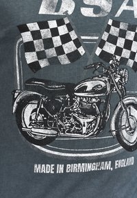 Sort-hvid grafik af en vintage motorcykel, sat mod en mørkegrå baggrund, med ternede flag og tekst, der lyder "LAVET I BIRMINGHAM, ENGLAND."