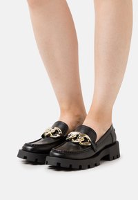 Schwarze, klobige Slipper mit großen silbernen Ketten-Details, die barfuß vor einem schlichten weißen Hintergrund getragen werden.