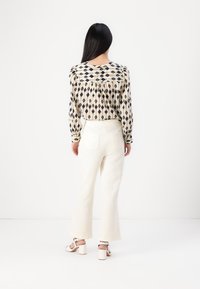 Långärmad blus i beige med marinblå diamantmönster, ihopparad med vita texturerade vida byxor och vita sandaler med klack.
