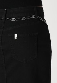 Zwarte denim broek met een zilveren kettingaccent rond de taille, voorzien van een klein zilveren merklabel op de achterzak. Minimalistisch ontwerp.
