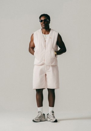 Mand iført lys lyserød ærmeløs vest og matchende shorts med hvide sokker, sølvfarvede sneakers, solbriller og gyldt ur, stående foran ensfarvet baggrund.