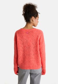 Korallenfarbener Strickpullover mit strukturiertem Muster, gerippten Bündchen und Saum, mit einem kleinen rosa Akzentknopf auf der Rückseite. Kombiniert mit grauen Hosen.