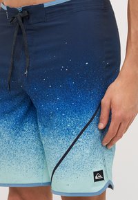 Costum de baie cu un gradient albastru și un design pistruiat, cu un brâu ajustabil, accente pe lateral și un mic logo brodat pe tiv.