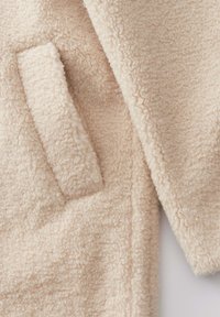 Cappotto in pile beige con una superficie morbida e testurizzata. Presenta una tasca frontale e un design dal taglio morbido. Ideale per calore e comfort.