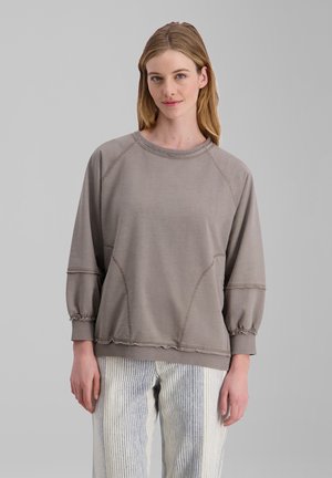 Junge Frau mit hellbraunen Haaren trägt ein lockeres graues Sweatshirt und weiße Hose mit vertikalen blauen Streifen vor einem einfarbigen grauen Hintergrund.