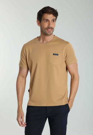 Homme portant un t-shirt beige à manches courtes avec une poche poitrine portant l'inscription "EST.90" et un pantalon noir, debout avec une main dans la poche.