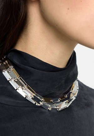 Błyszczący srebrny i złoty prostokątny łańcuch z ogniw noszony na czarnym golfie na szyi osoby.