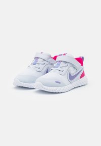 Zapatillas deportivas blancas con parte superior de malla, acentos morados y un forro rosa. Presenta una correa de gancho y bucle y una suela blanca texturizada.