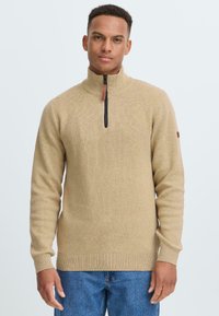 Beige gebreide trui met een hoge kraag en zwarte rits, voorzien van geribbelde manchetten en onderrand. Zachte textuur met een subtiel patroon.