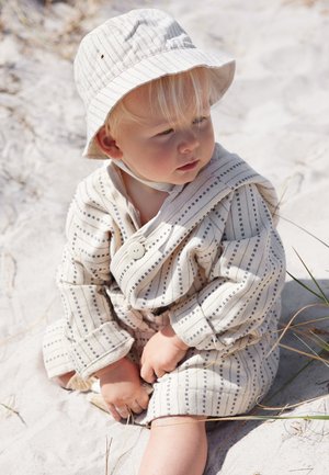Toddler wearing a striped jacket and sun hat, sitting on white sandy ground with sparse grass in bright daylight.