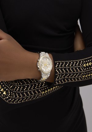 Wrist with silver and gold jeweled watch on arm crossed over black long sleeve shirt adorned with gold studs and triangle patterns.