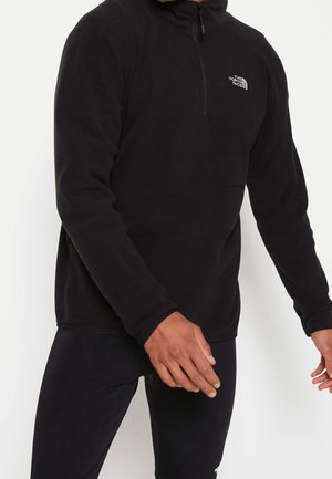 Mężczyzna ubrany w czarną polarową bluzę z długim rękawem i półzamek, z logo The North Face na piersi oraz pasujące czarne spodnie na jednolitym tle.