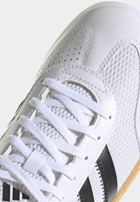 Chaussure de sport blanche avec tige en maille et cuir, lacets texturés, accents noirs et semelle extérieure en caoutchouc. Présente des sections perforées pour la respirabilité.