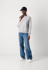 Felpa grigia con polsini a costine, abbinata a jeans a gamba larga blu. Sneakers bianche completano l'outfit. Un cappellino nero presenta del testo.
