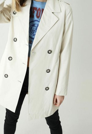 Personne portant un manteau croisé beige clair avec des boutons noirs sur un T-shirt graphique bleu et un pantalon noir, posant devant un fond uni.