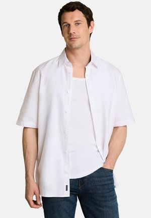 Mann mit kurzen dunklen Haaren, der ein offenes kurzärmeliges weißes Hemd über einem weißen Tanktop und blauen Jeans trägt, mit einer Hand in der Tasche.