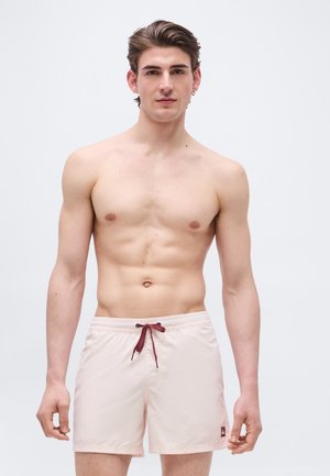 Mlad moški s kratkimi lasmi, oblečen v svetlo bež kopalke z bordo rdečo vrvico, stoji pred enostavnim svetlo sivim ozadjem.